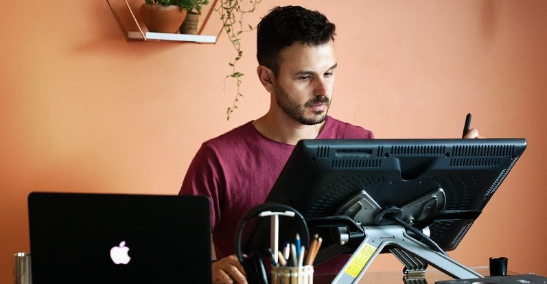 Alex Cói desenhando em uma mesa digitalizadora no estúdio, ao lado de um notebook.